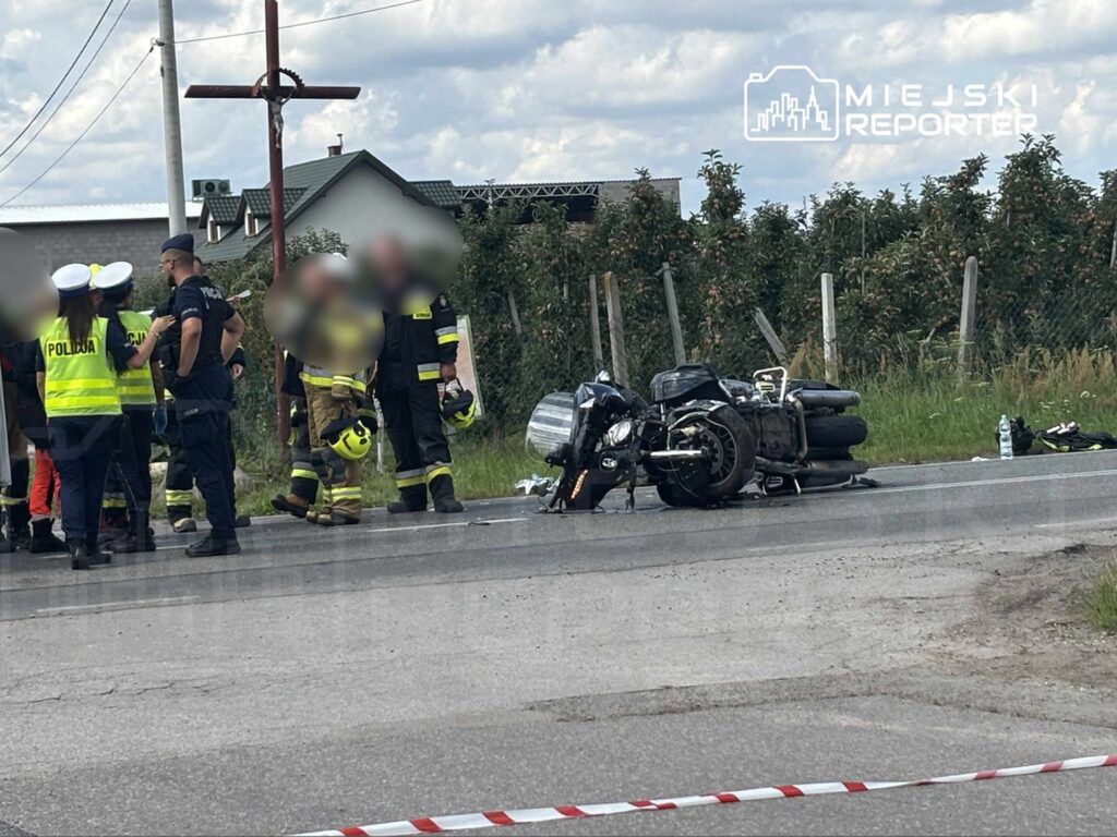 Grupa ratowników i policjantów pracuje przy przewróconym motocyklu na drodze, w tle widać domy i krzyż.