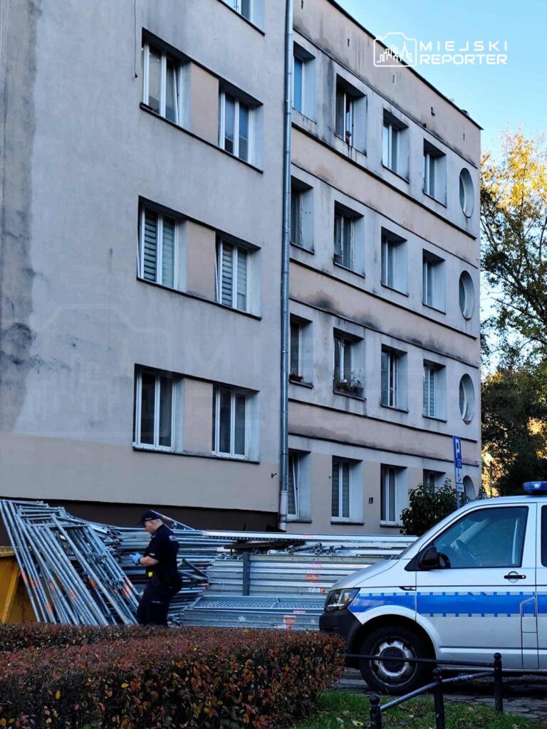 Policjant przeszukuje teren obok stosu rusztowań przy budynku mieszkalnym w miejskim otoczeniu.