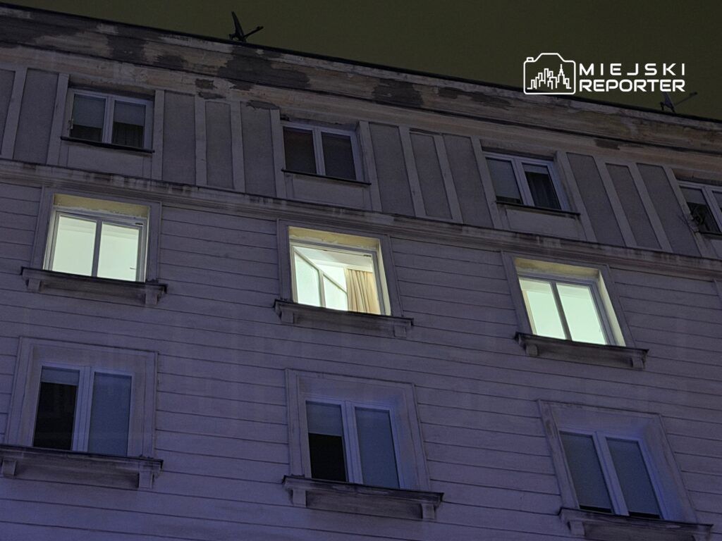 Oświetlone okno z zasłonami w budynku mieszkalnym nocą, w otoczeniu ciemnej elewacji.