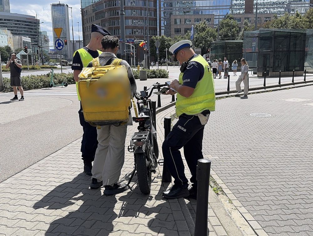 Trzech policjantów rozmawia z mężczyzną na rowerze dostawczym w miejskim otoczeniu, obok nowoczesnych budynków.