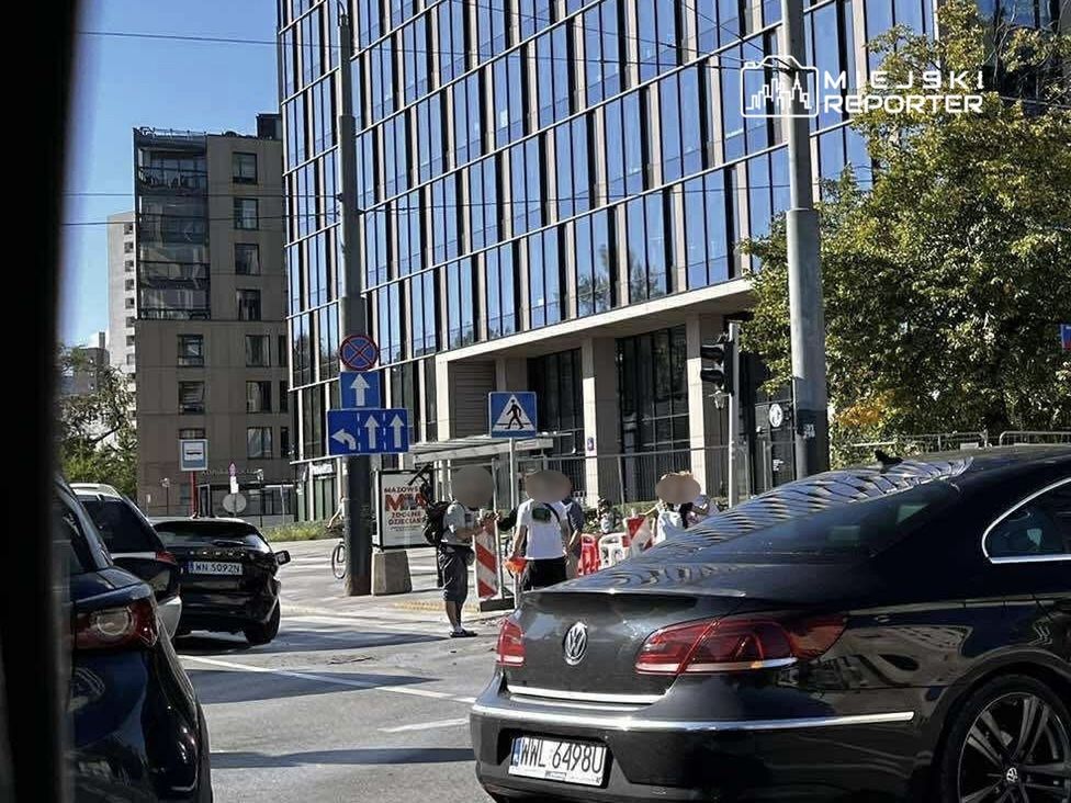 Grupa osób przechodzi przez ulicę w pobliżu nowoczesnego biurowca z przeszkloną fasadą, w tle znaki drogowe.