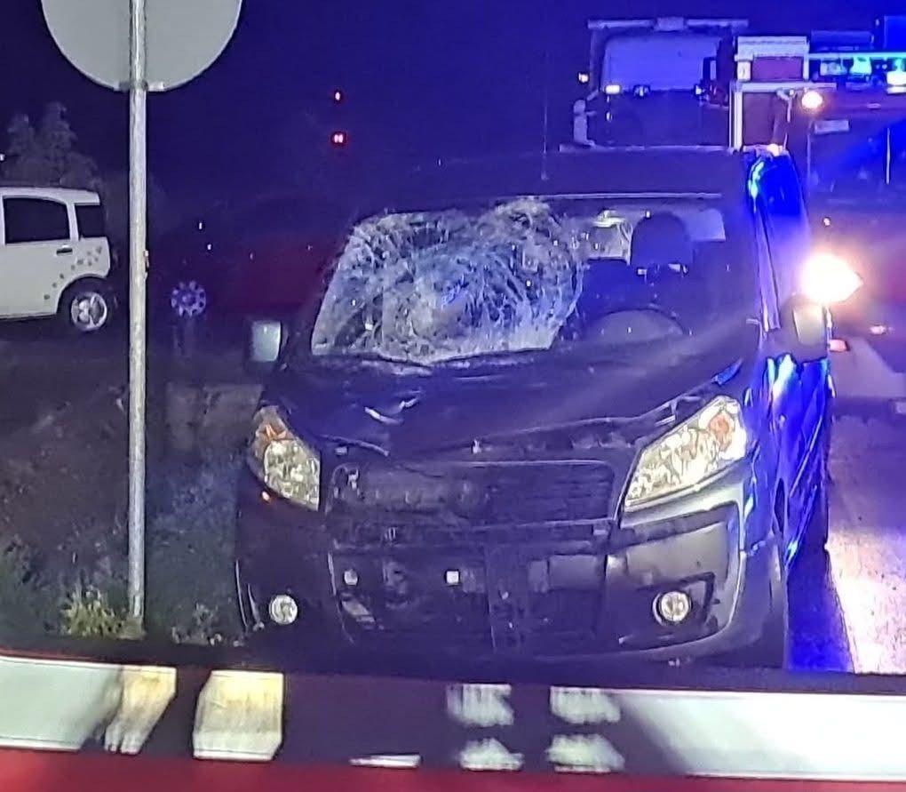 Rozbity czarny van z uszkodzoną przednią szybą stoi na poboczu drogi, obok znaku drogowym. W tle widać niebieskie światła pojazdów służb ratunkowych.