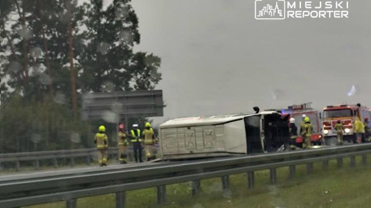 Zespół ratunkowy w żółtych hełmach pracuje przy przewróconym pojeździe na poboczu drogi, w tle widać straż pożarną.