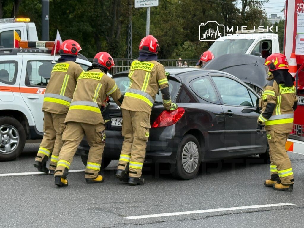 Grupa strażaków w mundurach, w kaskach, pcha uszkodzony samochód na drodze, obok stoi pojazd służb ratunkowych.