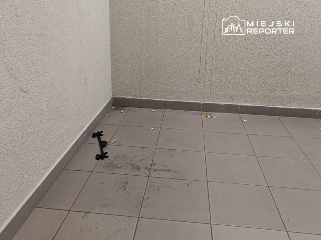 Pusty korytarz z brudną posadzką, na której leży czarny statyw i resztki śmieci. Ściany gładkie, jasne.