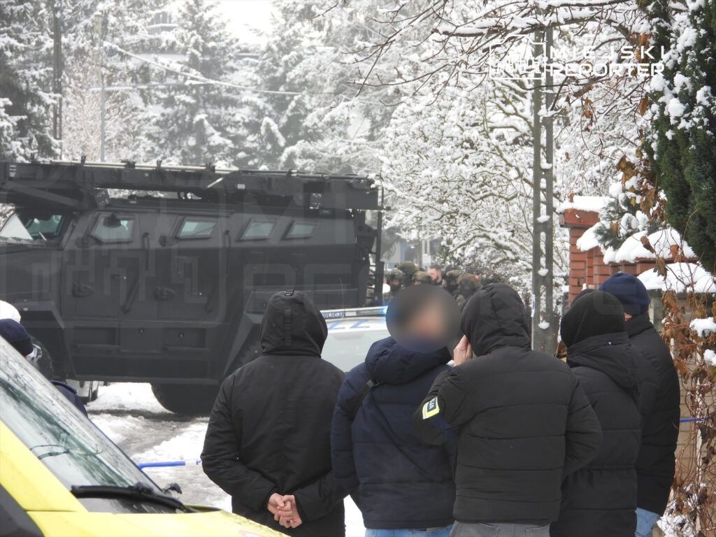 Grupa osób w zimowych kurtkach obserwuje akcję policyjną w zaśnieżonym otoczeniu, obok stoi opancerzony pojazd.