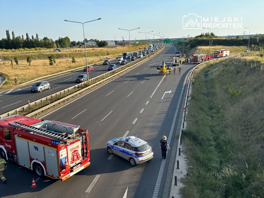 Na drodze krajowej widać straż pożarną oraz policyjny radiowóz, a obok trwają działania ratunkowe.