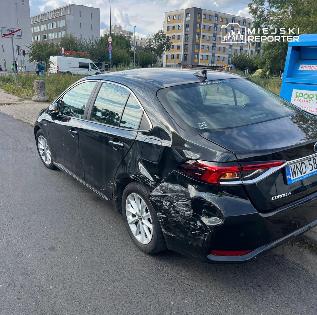 Czarny samochód osobowy z uszkodzeniami z lewej strony stoi na ulicy w miejskim otoczeniu.