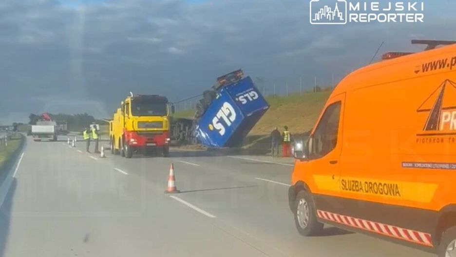 Czerwono-żółta ciężarówka z podnośnikiem stabilizuje przewróconą niebieską ciężarówkę na autostradzie.