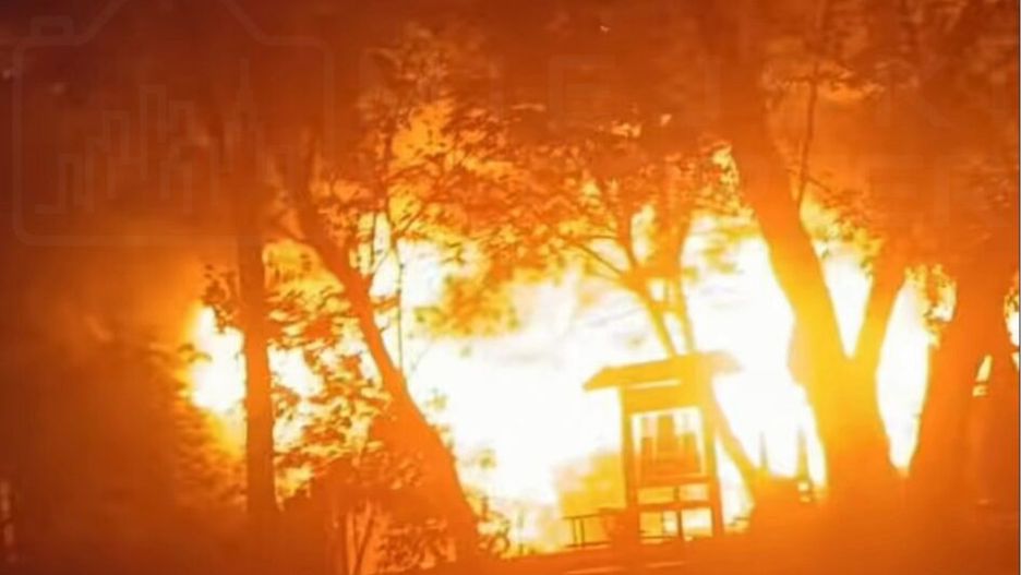 Ognisko pożaru w lesie, otoczone drzewami, z intensywnym płomieniem i dymem w tle.