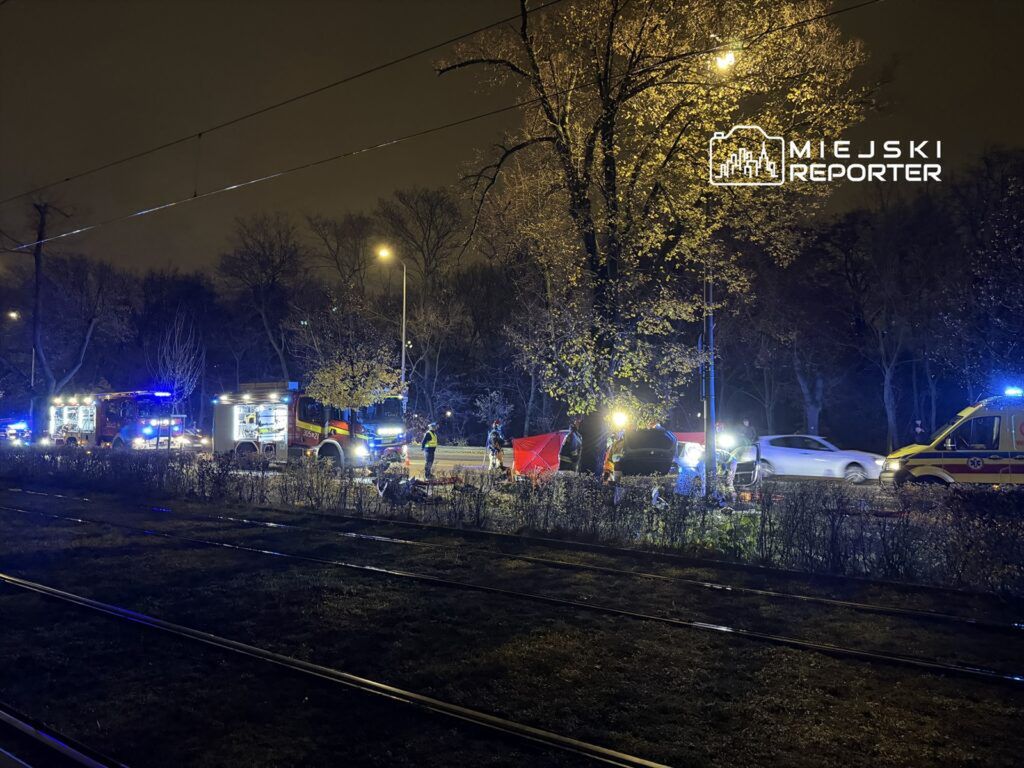 Strażacy oraz ratownicy medyczni pracują na miejscu wypadku drogowym w nocy, otoczeni przez pojazdy służb ratunkowych.