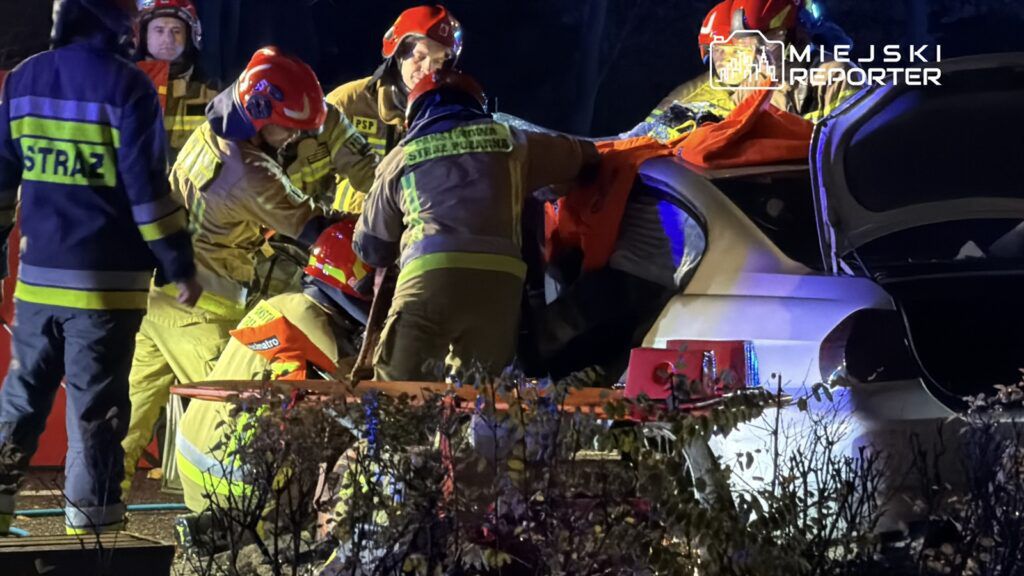 Grupa strażaków w specjalistycznym ubraniu prowadzi akcję ratunkową przy uszkodzonym samochodzie w nocy.