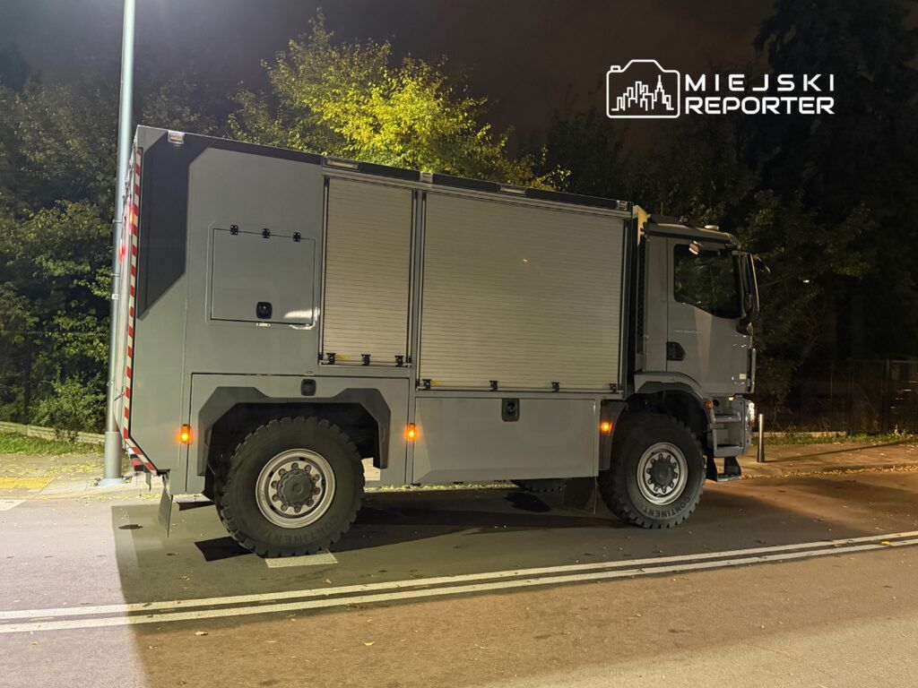 Szary pojazd specjalistyczny zaparkowany na ulicy w nocy, otoczony drzewami w tle.