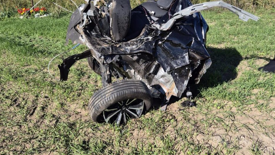 Zniszczony samochód leży na polu, obok krzyż z kwiatami, w tle zielone pola i niebo z chmurami.