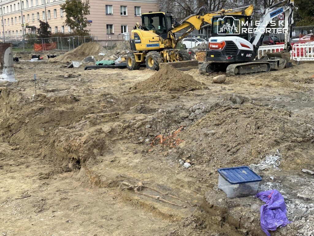Na placu budowy pracują maszyny budowlane, w tym koparki, w otoczeniu wykopów i materiałów budowlanych.