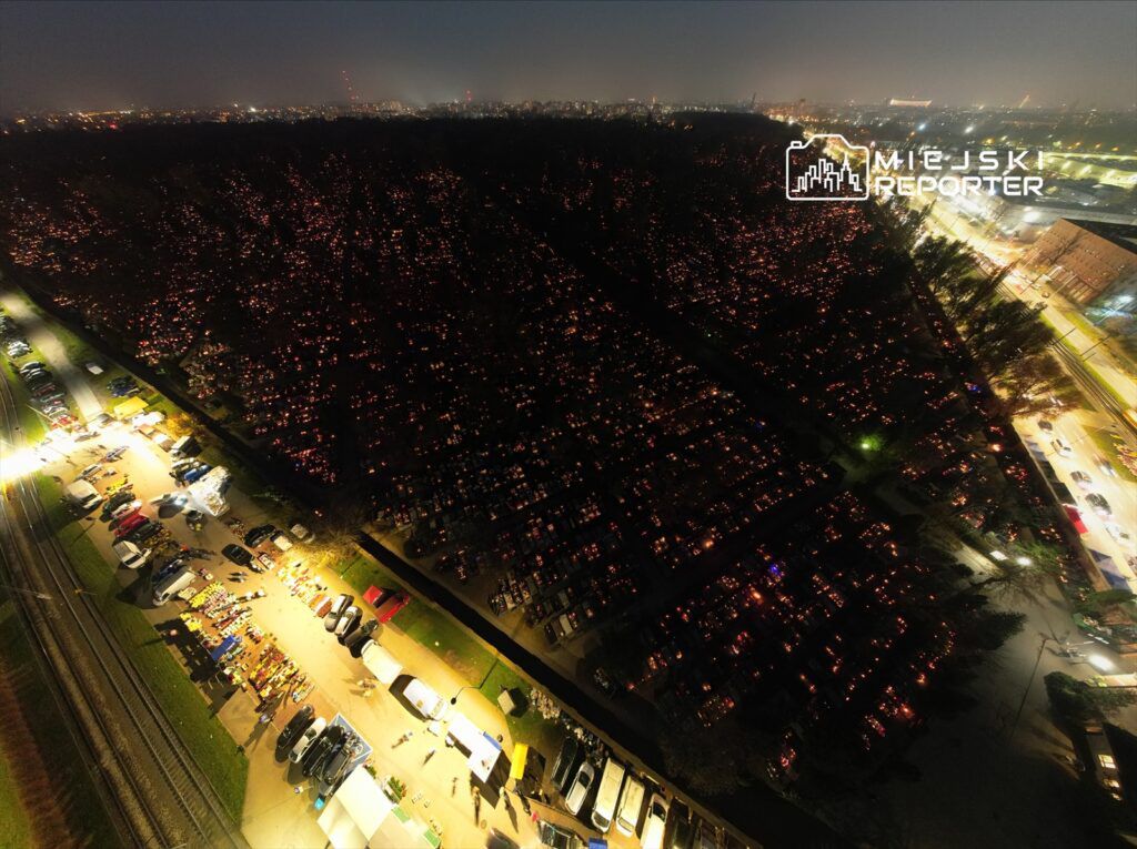 Widok z góry na cmentarz z setkami zapalonych zniczy w nocy, obok parking z samochodami i straganami.
