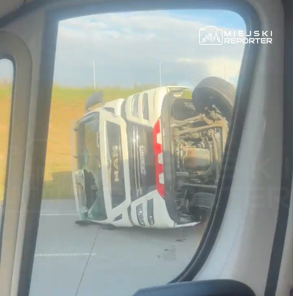 Przewrócony samochód ciężarowy leży na boku na drodze, otoczony zielonym krajobrazem.