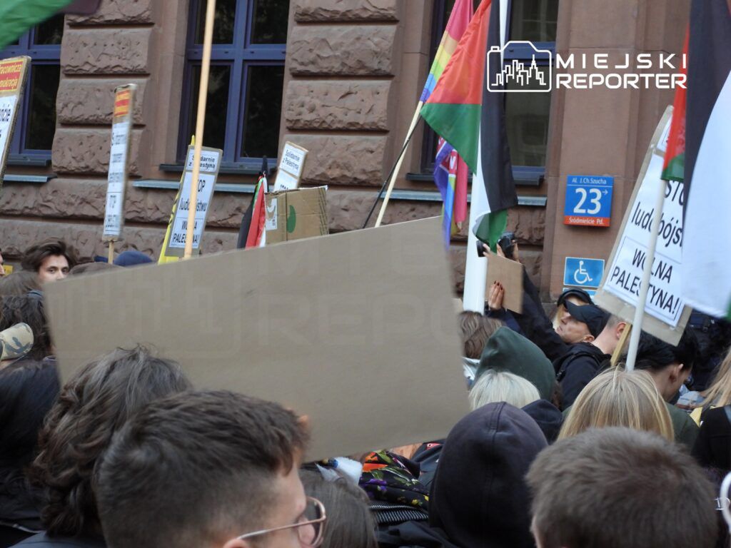 Tłum ludzi z transparentami i flagami, protestujących przed budynkiem z brązową elewacją.