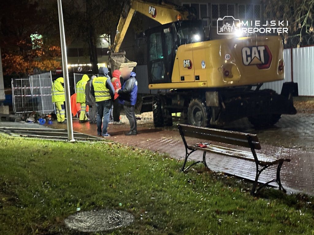 Grupa pracowników w odblaskowych kamizelkach rozmawia obok koparki na deszczowej nawierzchni w parku.