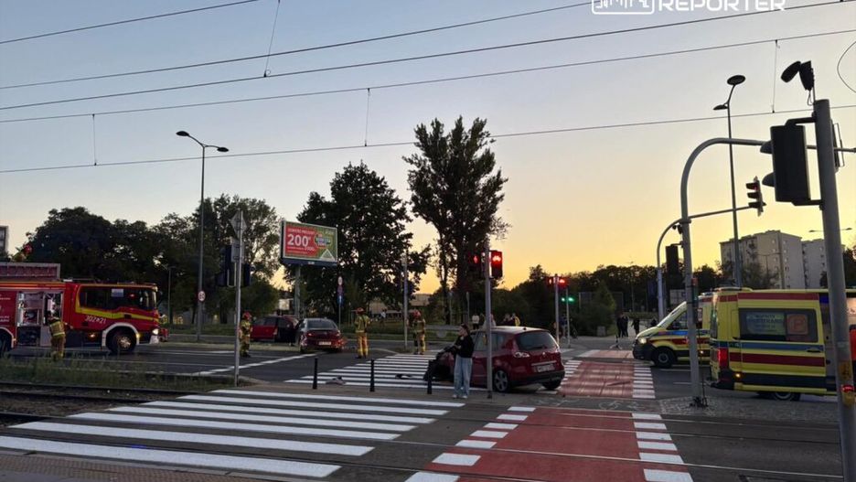 W miejscu zdarzenia drogowego, strażacy i ratownicy medyczni pracują przy uszkodzonym samochodzie na przejściu dla pieszych.