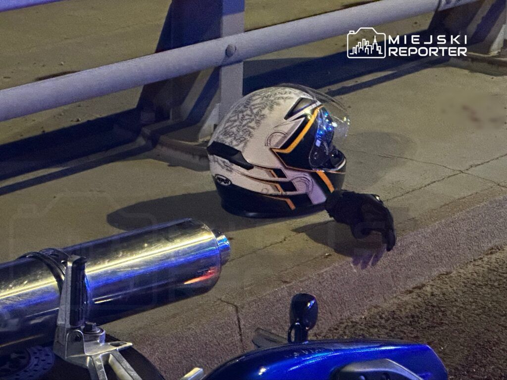Kask motocyklowy i rękawica leżące na chodniku obok metalowej rury wydechowej.