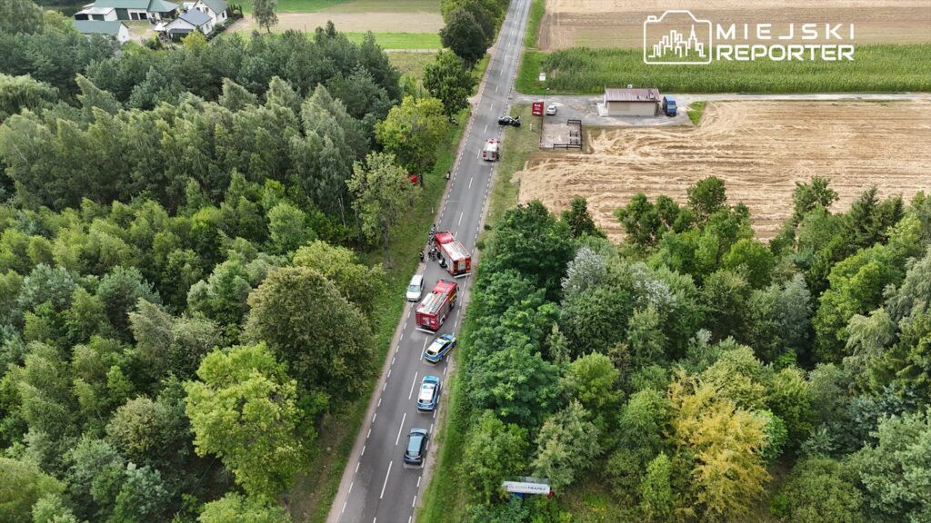 Na drodze w otoczeniu lasu i pól stoją pojazdy straży pożarnej oraz radiowozy, a obok widać zniszczony samochód.