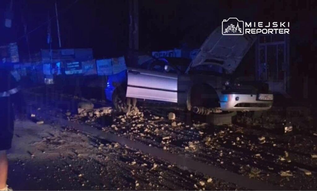 Samochód z otwartą maską stoi na drodze, obok niego leży gruz, a w tle widoczne są niebieskie światła policyjne.
