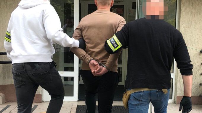 Mężczyzna w kajdankach prowadzony przez dwóch policjantów do wejścia do komisariatu.