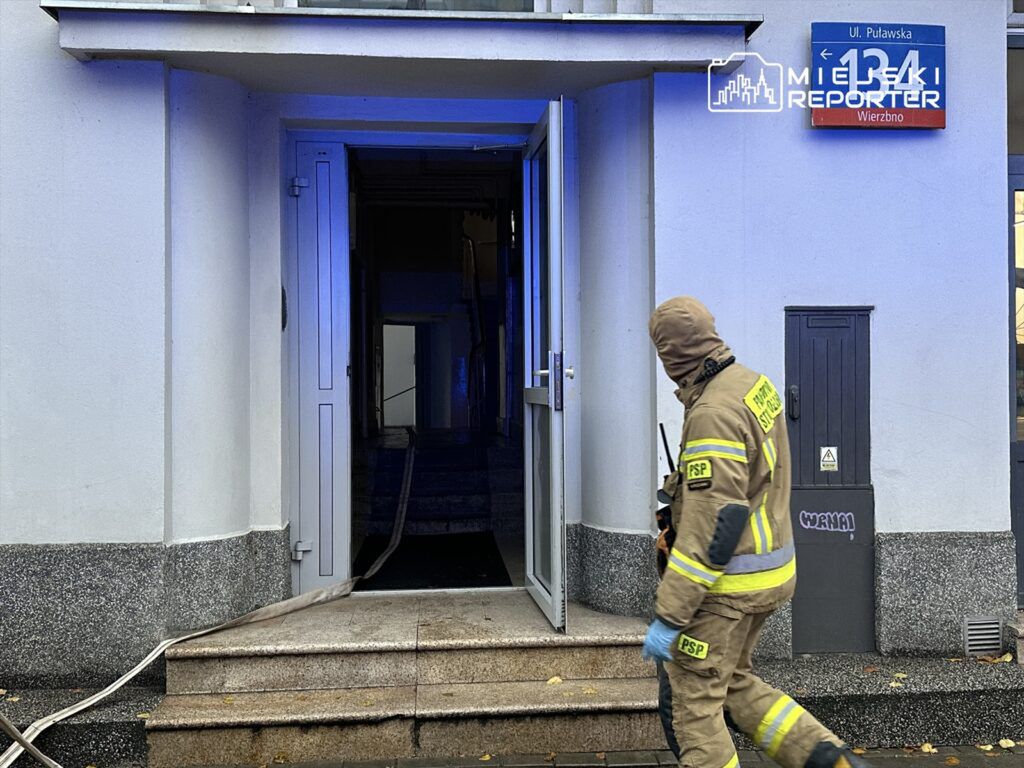 Strażak w specjalnym ubraniu przechodzi obok wejścia do budynku z wężem gaśniczym na ziemi.