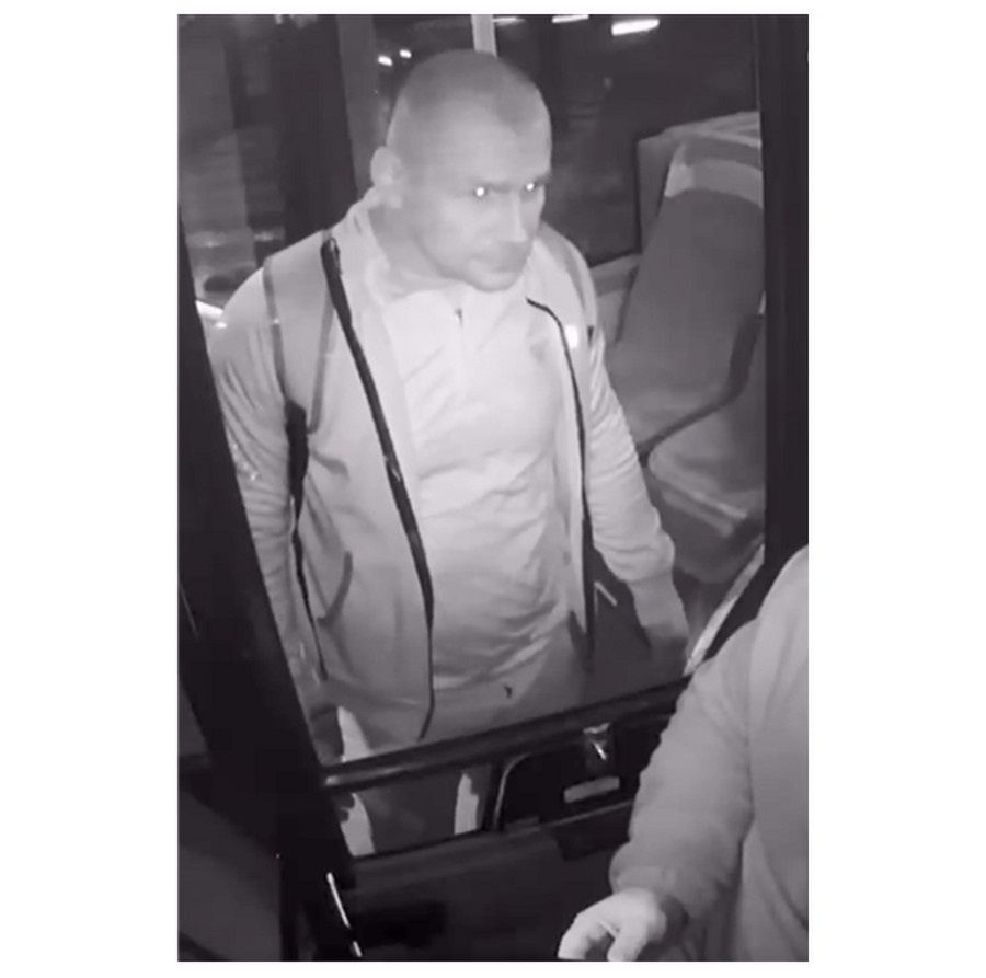 Mężczyzna w jasnym dresie wchodzi do autobusu, stojąc blisko drzwi, obok drugiego mężczyzny w ciemnym ubraniu.