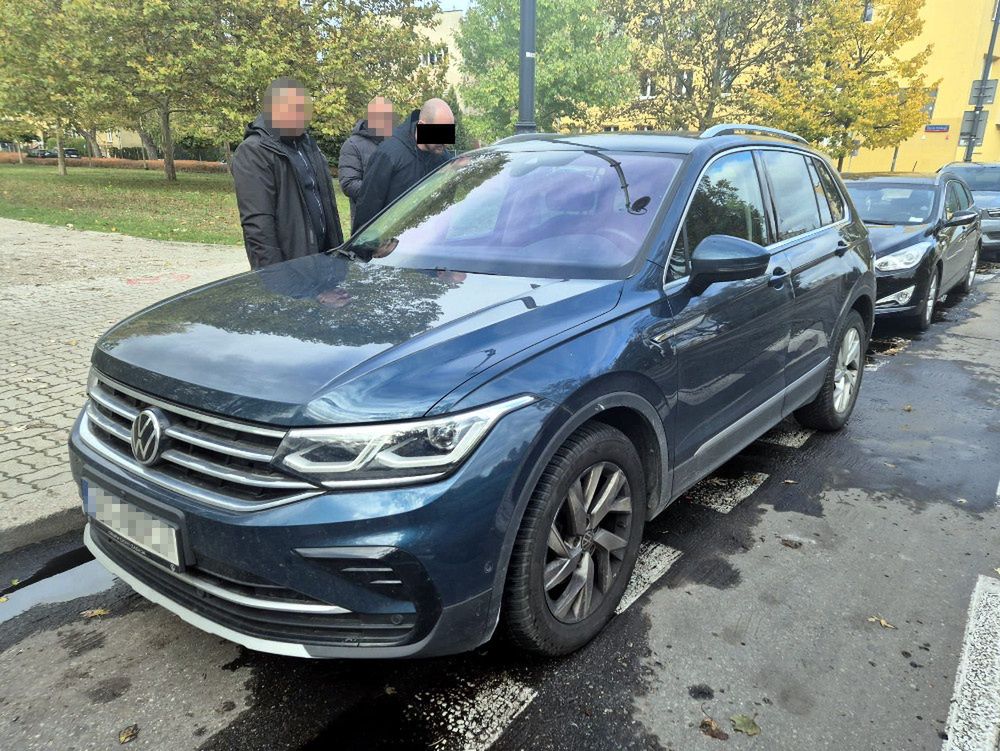 Trzech mężczyzn rozmawia obok niebieskiego samochodu marki Volkswagen zaparkowanego na ulicy, w tle drzewa.