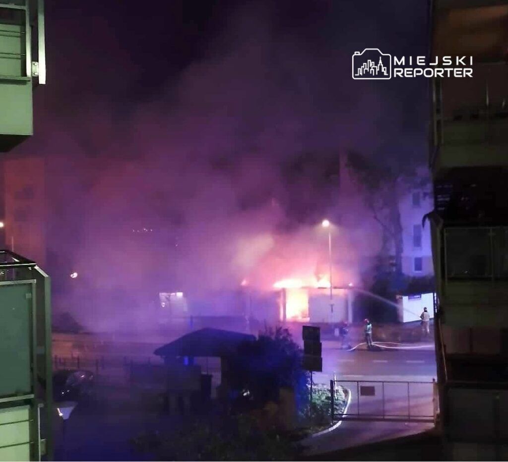 Płonący budynek otoczony gęstym dymem, w tle strażacy gaszący pożar w nocy.