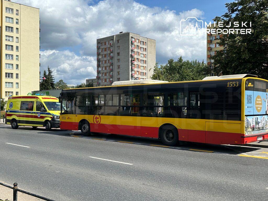 Karetka pogotowia zatrzymana obok miejskiego autobusu na ruchliwej ulicy, w tle bloki mieszkalne i niebo z chmurami.