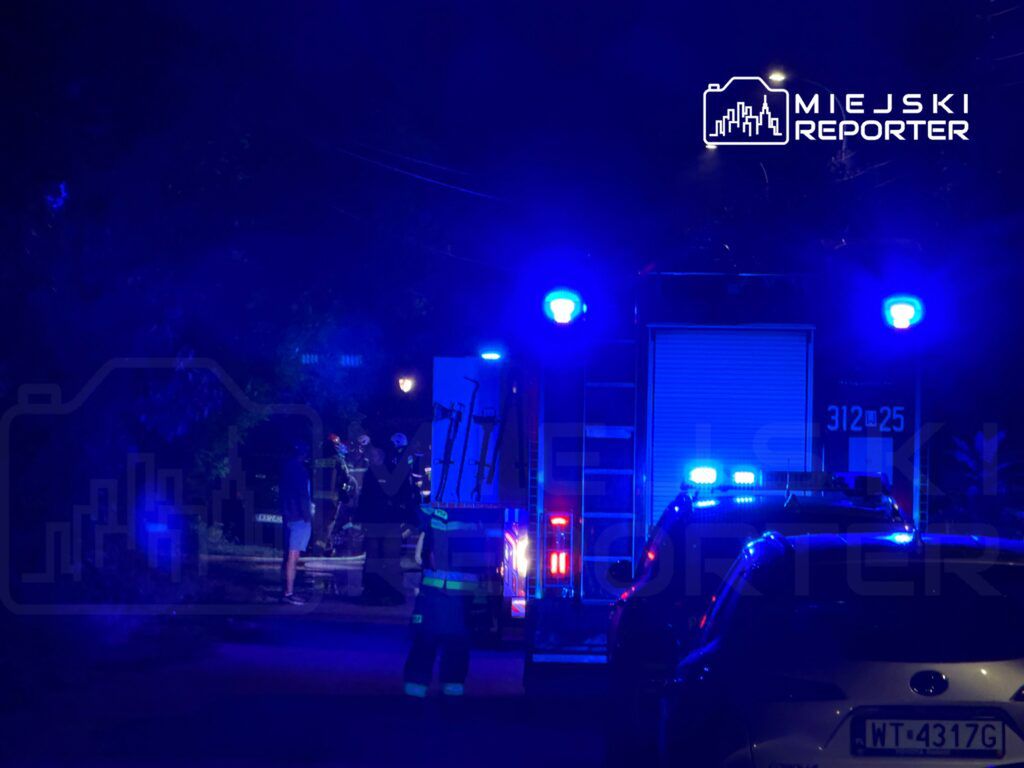 Grupa strażaków w mundurach z wężem gaśniczym, pracująca w ciemnej ulicy oświetlonej niebieskimi światłami.