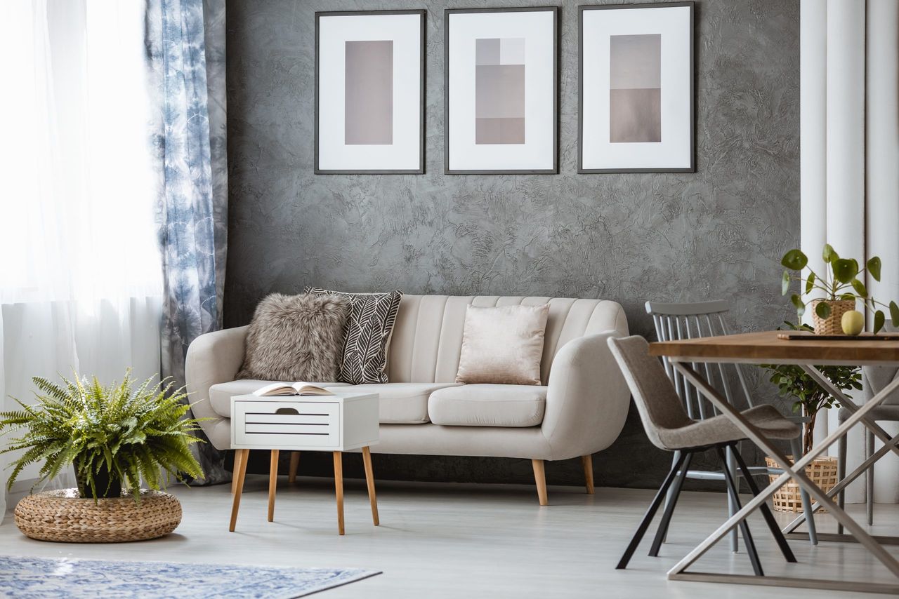 Simple living room with beige couch, white stool and fern on rattan pouf near the window