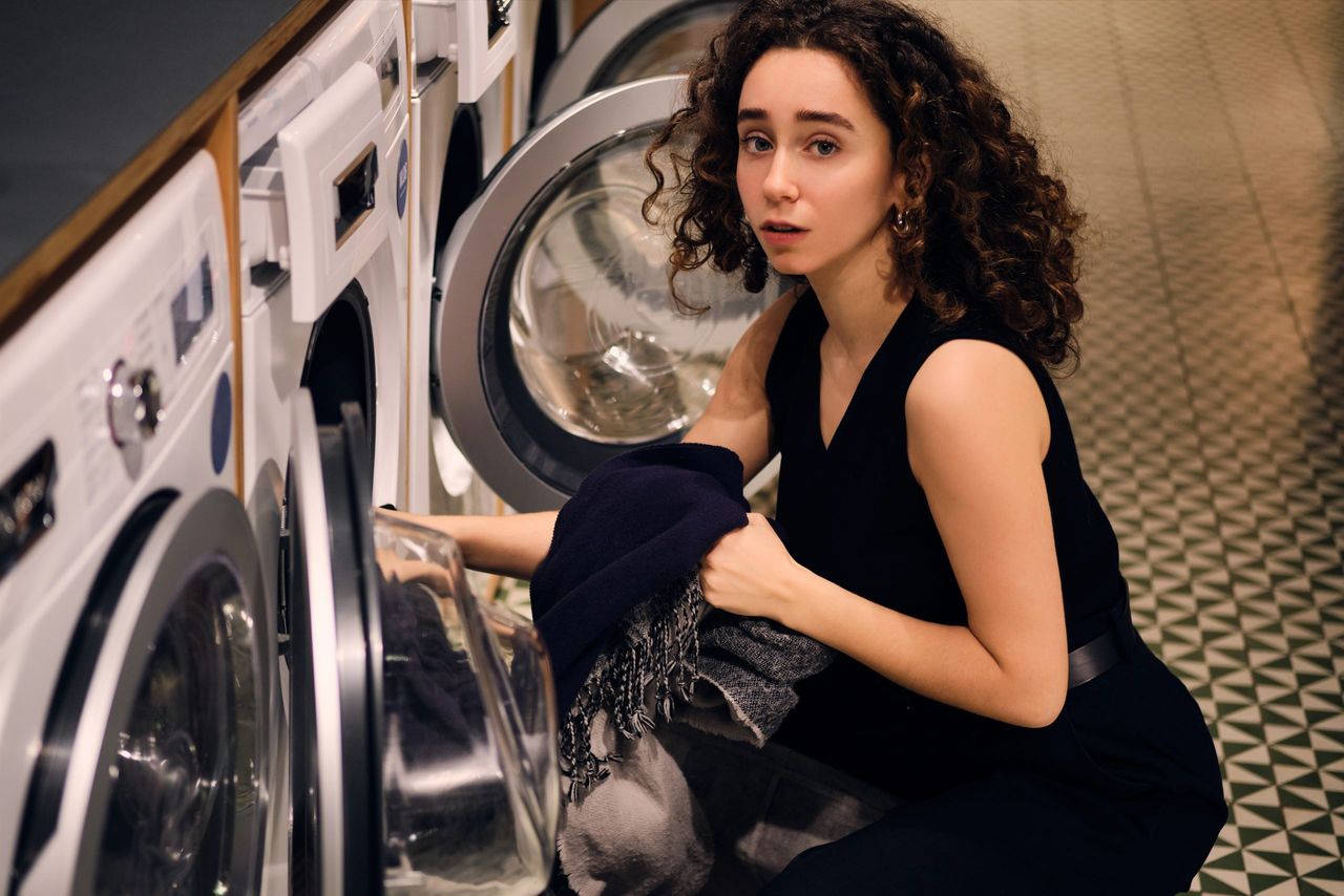 Portrait of brunette girl thoughtfully looking in camera taking out clothes from washing machine in self-service laundry