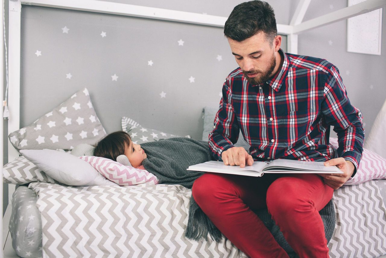 Father reading a book to his daughter while lying on the floor in bedroom.