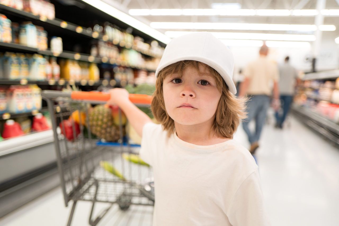 Healthy food for young family with kids. Boy at grocery store or supermarket. Portrait of funny little child holding shopping bag full of fresh vegetables. Child with shopping trolley with products