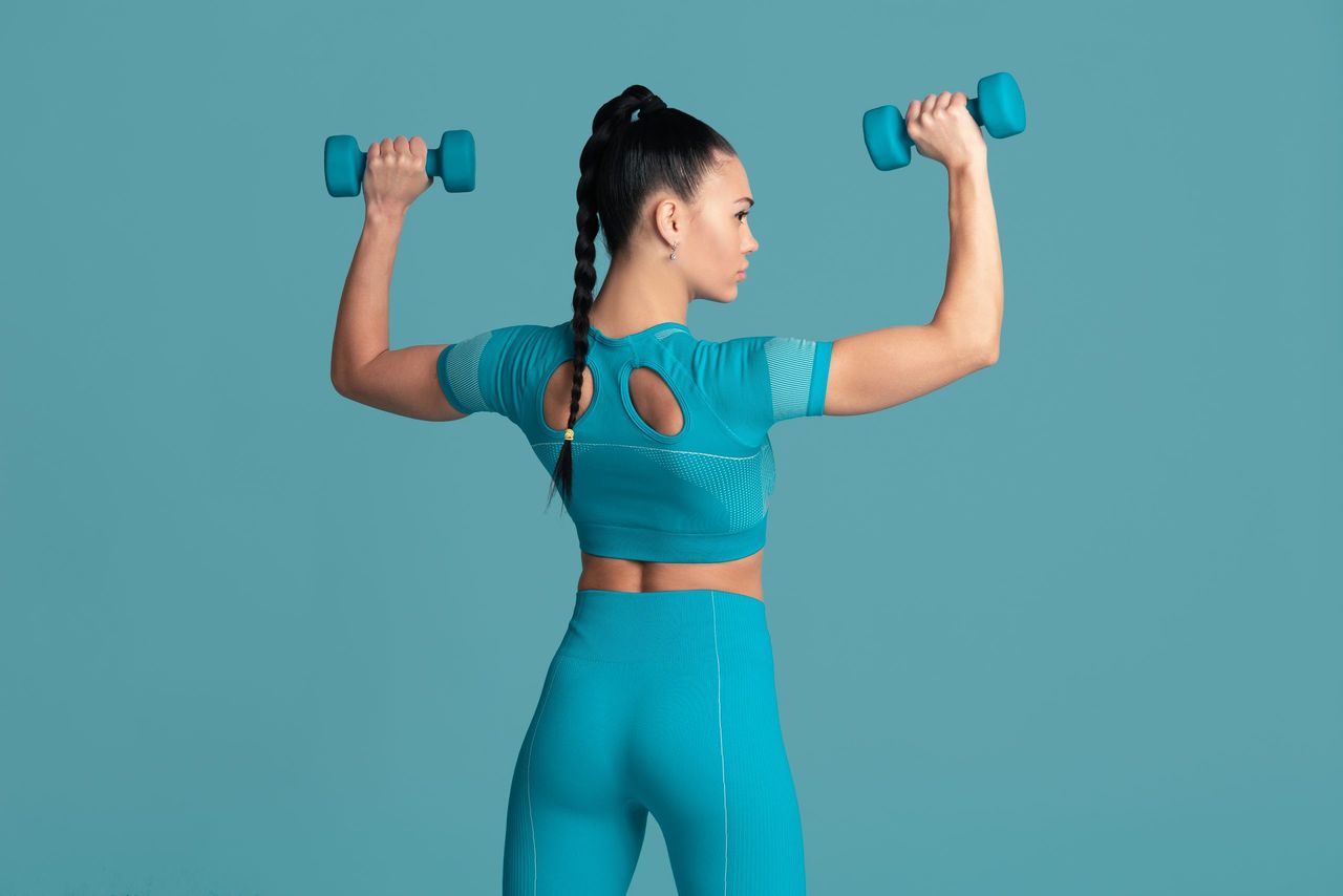 Confidence. Beautiful young female athlete practicing in studio, monochrome blue portrait. Sportive fit brunette model with weights. Body building, healthy lifestyle, beauty and action concept.