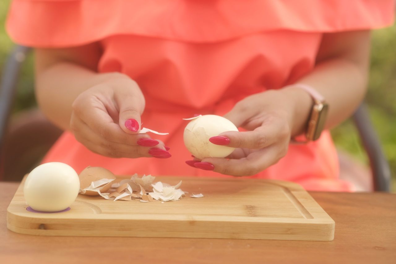 Unrecognizable lady peeling boiled egg on wooden board outdoors. Woman's hands with bright manicure peeling chicken egg from shell on wooden board. Close up