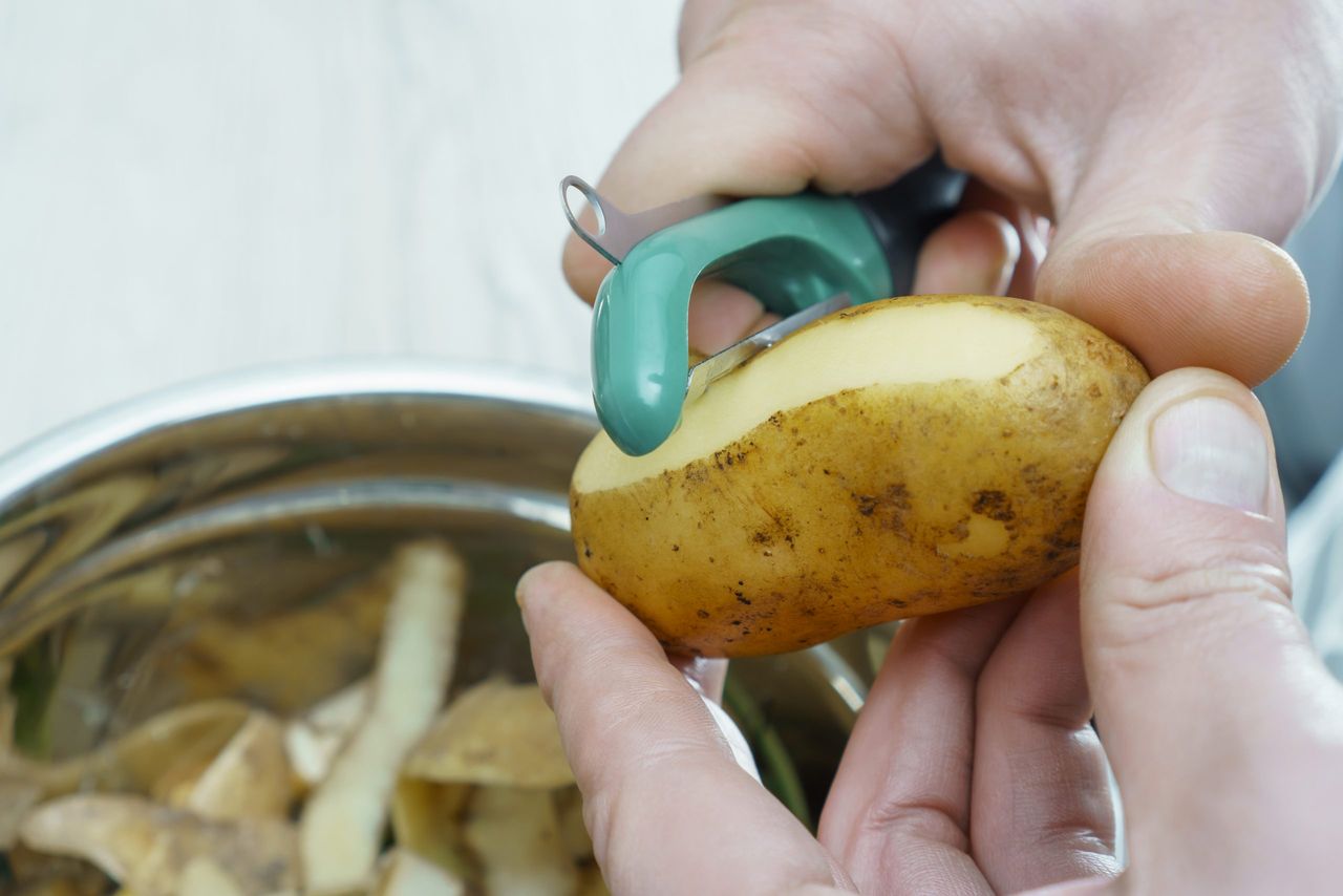 Close-up of unrecognizable man wearing peeling potato with peeler knife over metal bowl full of peelings, standing in kitchen. Recycling, composting, fertilizing, zero waste, organic waste management.