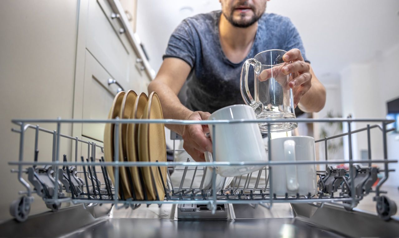 A man in front of an open dishwasher takes out or puts down dishes.