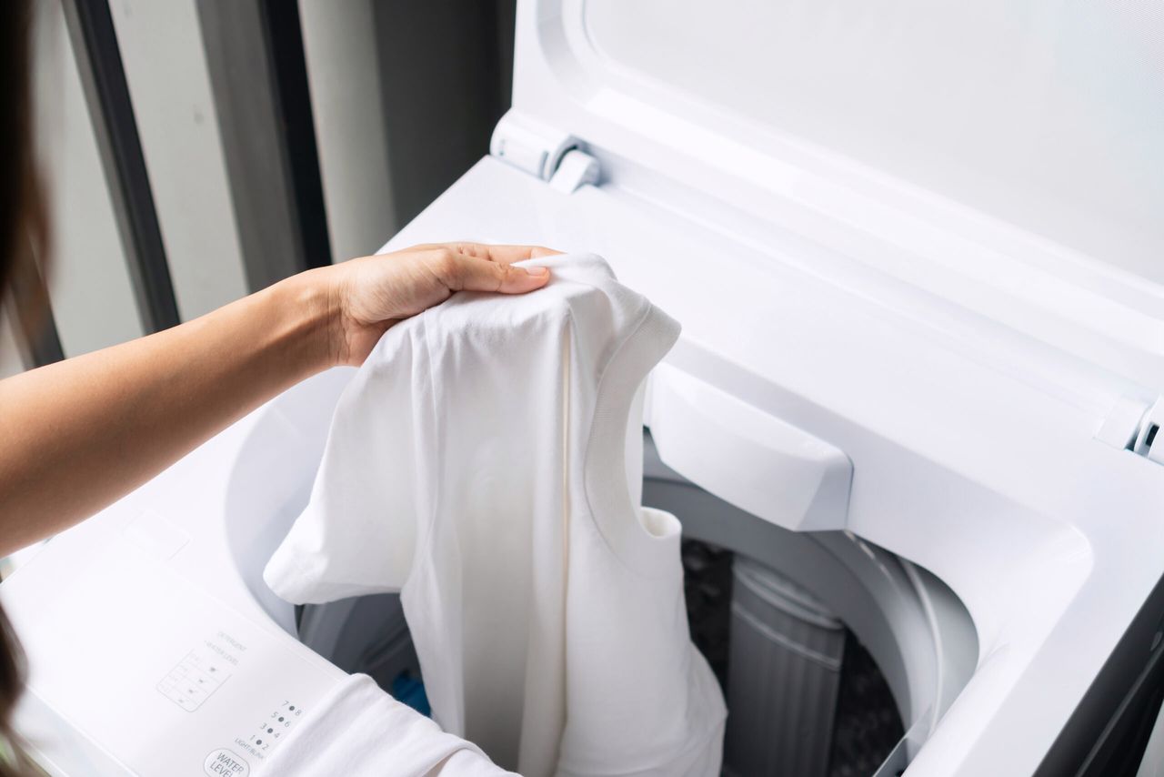 Hnads of young woman putting white color clothes into washing machine at condominium. Laundry concept. Top view