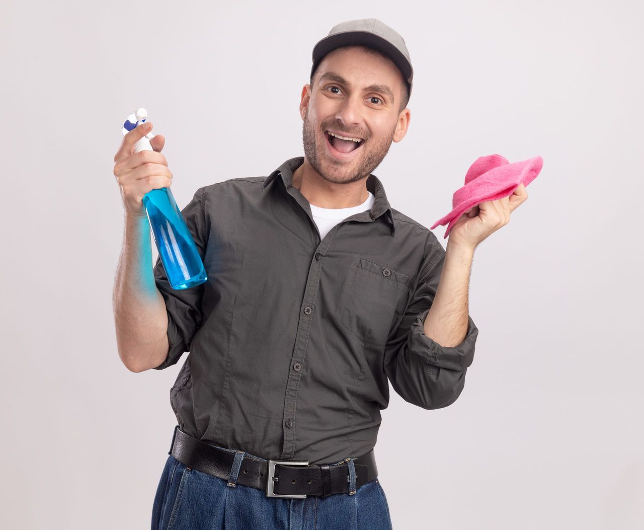joyful young cleaning man wearing casual clothes and cap holding cleaning spray and rag looking at camera happy and excited standing over white background