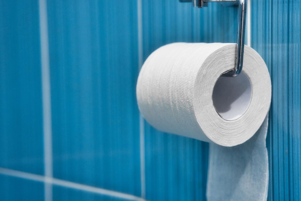 A roll of toilet paper hangs on a metal holder against a blue tile wall. Part of the toilet with space to copy