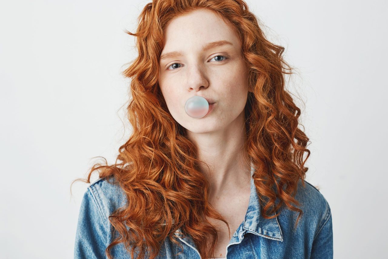 Cool brutal redhead girl in jean jacket chewing gum looking at camera over white background.