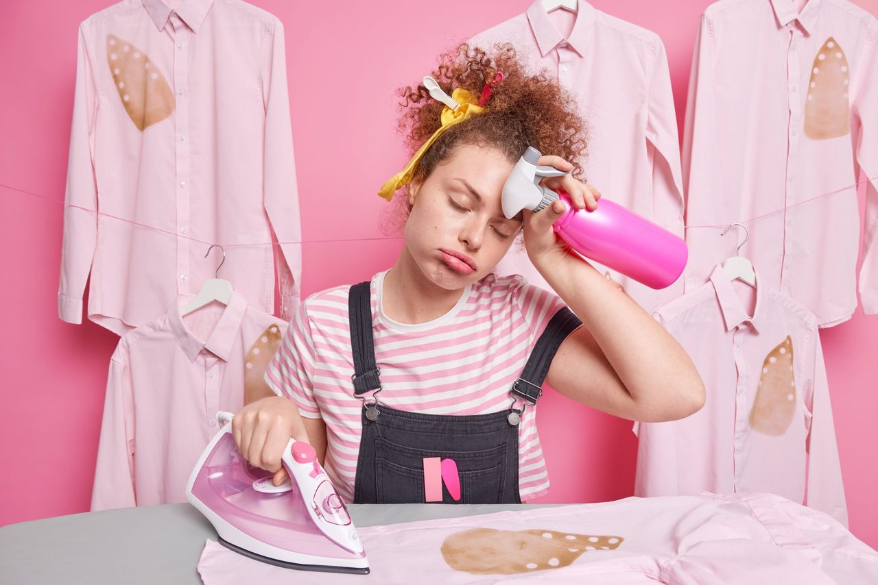 An overworked, sleepy woman housekeeper wipes her forehead, feeling fatigue while doing housework, ironing clothes, doing domestic chores, holding a spray bottle, and dealing with a burned shirt, all in a hurry to finish everything.