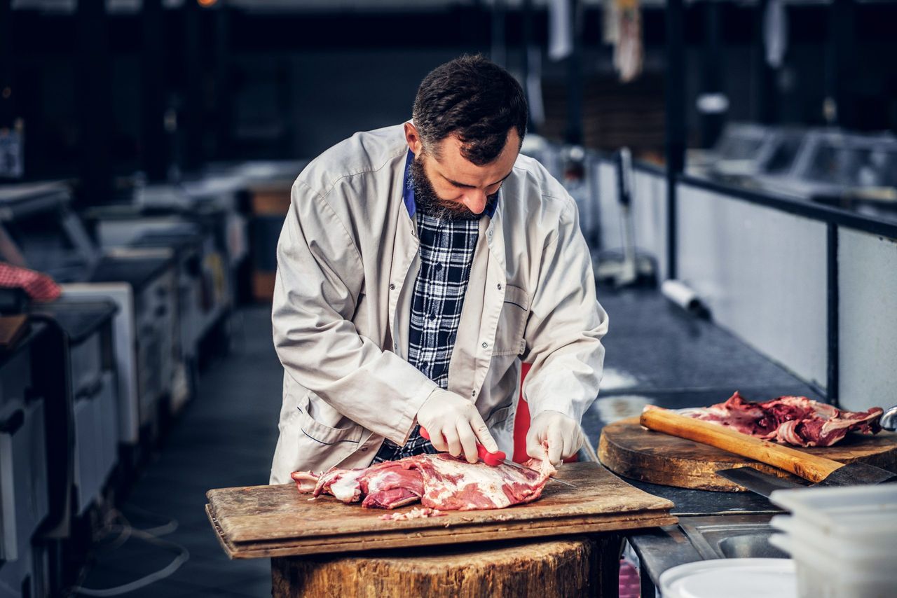 Bearded butcher dressed in a fleece shirt cut fresh bacon meat.