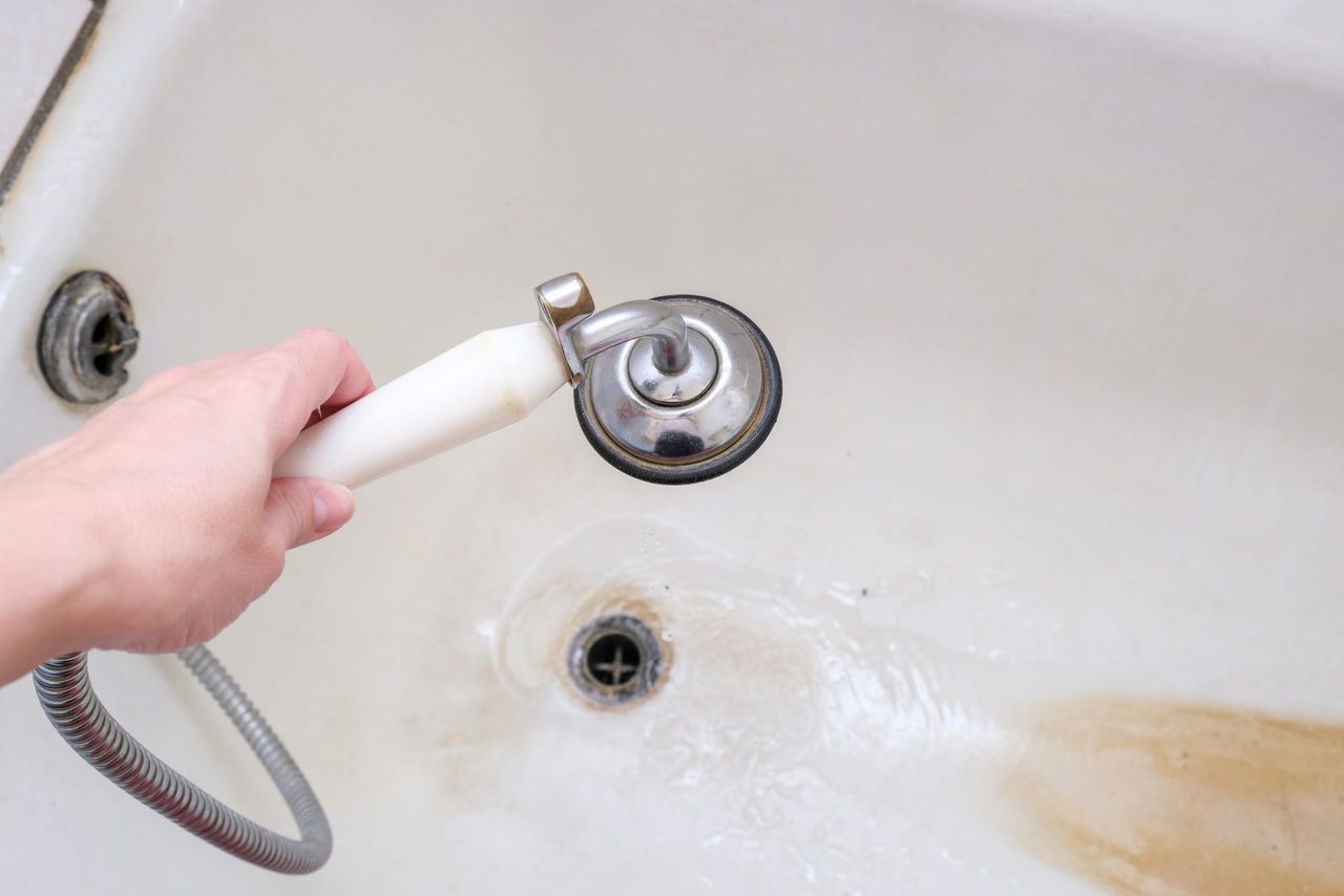 A hand holding shower head with running water against rusty bathtube with limescale, bathroom clining and decalcifying.