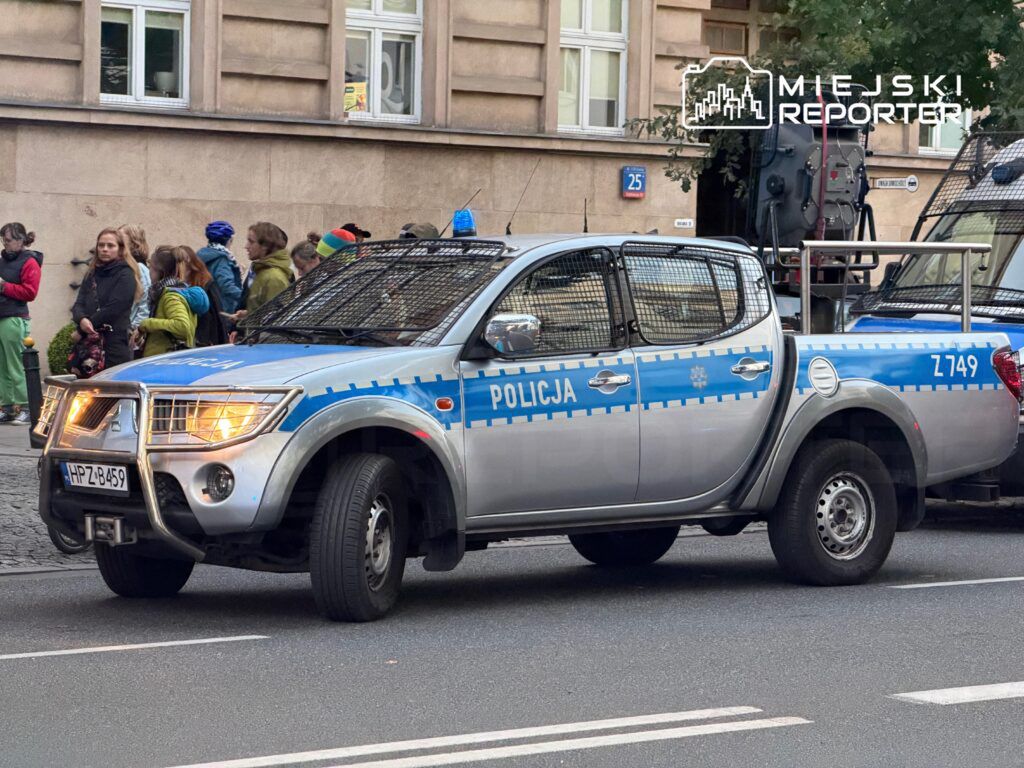 Radiowóz policyjny z niebieskimi oznaczeniami stoi na ulicy, obok grupy ludzi obserwujących okolicę.